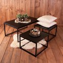 Ensemble de tables basses noires avec une grande et deux petites tables sur un plancher en bois, avec des plantes, des coussins et une peau de mouton.