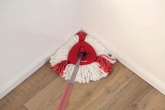 Serpillère rotative Vileda aux franges rouges et blanches dans le coin d'une pièce avec un parquet.