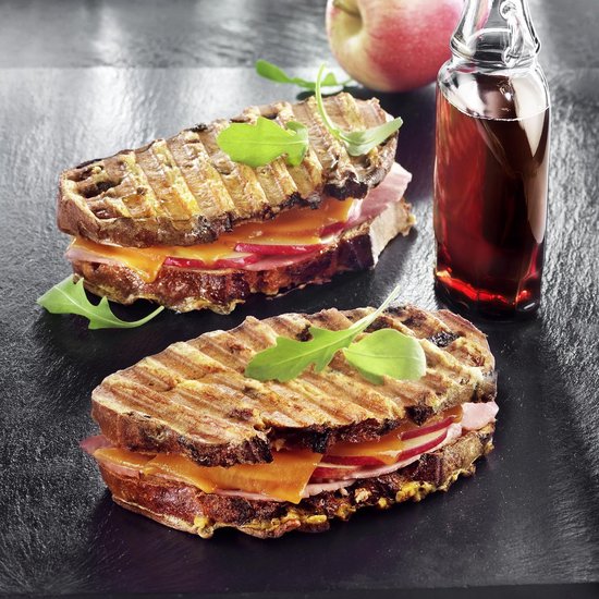 Twee gegrilde tosti's met ham, kaas, radijs en rucola, naast een appel en glas bessensap.