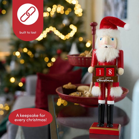 Houten kerstman notenkraker op een glazen tafel met een kerstboom op de achtergrond. Een beker en een dienblad met koekjes zijn ook te zien.