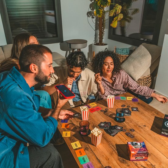 Drie vrienden spelen Hitster aan een tafel met de 'Movies & TV Soundtracks' doos, speelkaarten en popcorn.