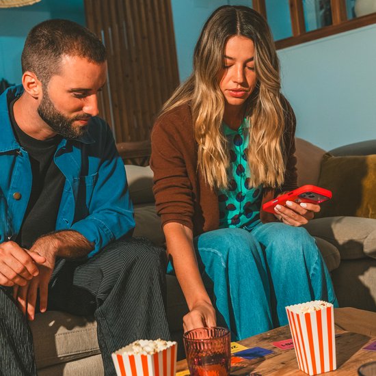 Een man en vrouw spelen Hitster aan tafel; de vrouw scant een kaart met haar smartphone, met popcorn binnen handbereik.