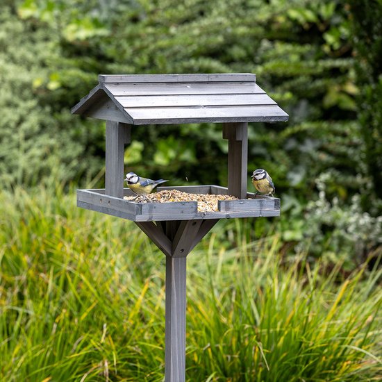 Grijs houten vogelvoederhuisje op paal met twee pimpelmezen die vogelzaad eten in een tuin.