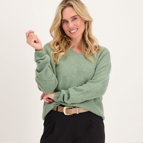 Model draagt een saliegroene trui met V-hals, zwarte broek en beige riem.