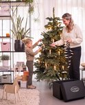 Vrouw en kind versieren een realistische kerstboom met kaarsverlichting, naast een doos met 'Christmas 1967' logo.