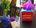 Een woonkamer met een boeket tulpen, ingepakte cadeaus met blauwe en paarse sjaals, en een rood speelgoedkistje met 'What's Inside?' erop.