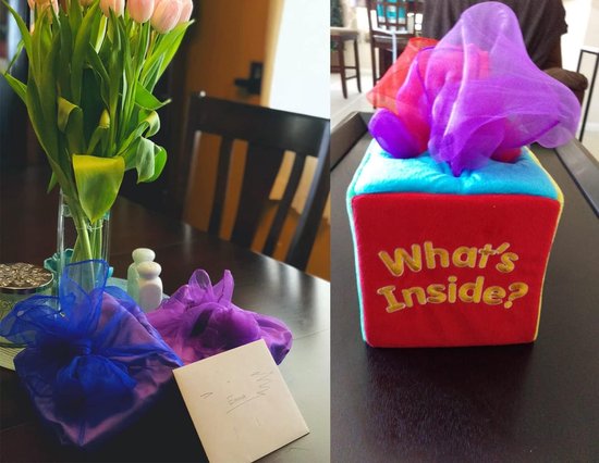 Een woonkamer met een boeket tulpen, ingepakte cadeaus met blauwe en paarse sjaals, en een rood speelgoedkistje met 'What's Inside?' erop.