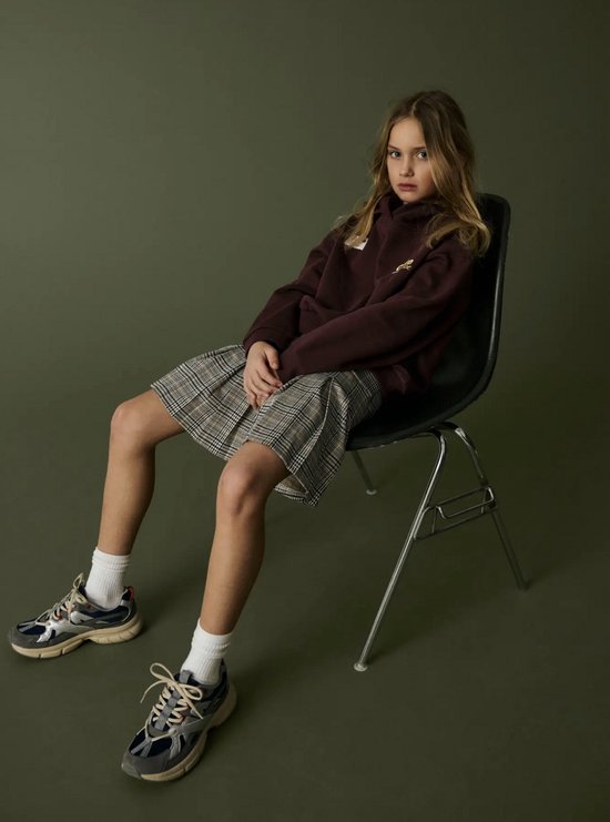 Meisje draagt een geplooide rok met ruitpatroon, bordeaux hoodie en sportieve sneakers.