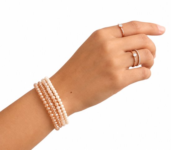 Vrouwenarm met vier kralenarmbanden in beige en roze tinten en twee verfijnde ringen met steentjes.