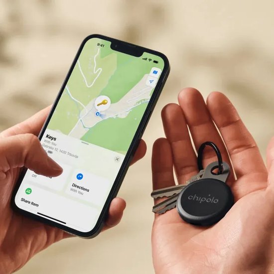 Een hand houdt een smartphone met de 'Find My'-app, terwijl de andere hand een zwarte Chipolo-tracker aan sleutels houdt.