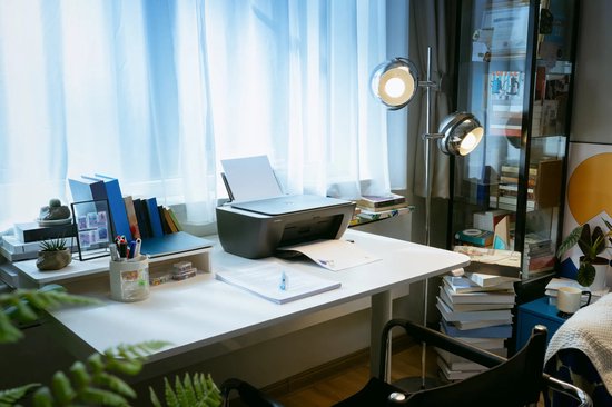 Zwarte HP DeskJet printer op een wit bureau bij een raam, met geprint document en boeken in de buurt.