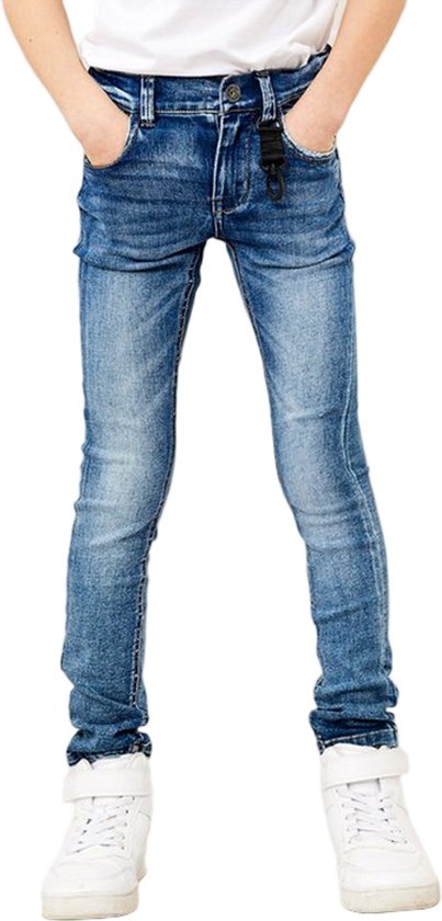 Jong kind met handen in zakken, draagt een skinny spijkerbroek met blauwe wassing en witte sneakers.