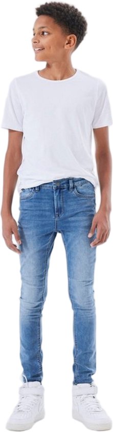 Jongen met gekruld haar, draagt een witte t-shirt en een skinny spijkerbroek met blauwe wassing.
