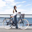 Vrouw in witte blouse loopt naast een zwarte RCB-fiets met mand op een kade langs het water.