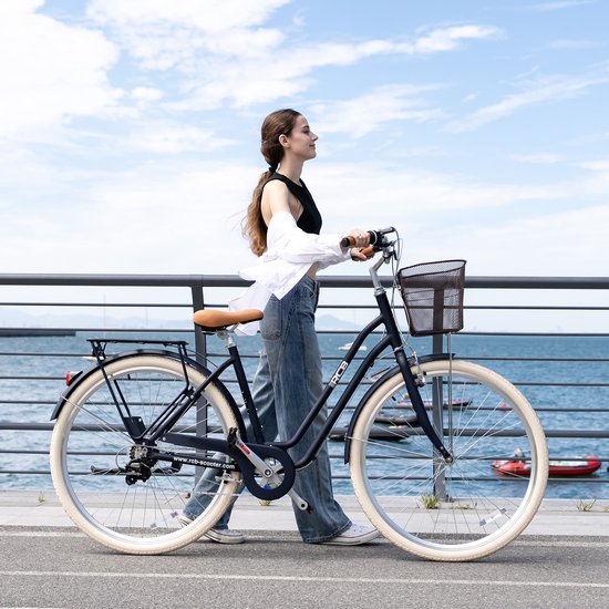 Vrouw in witte blouse loopt naast een zwarte RCB-fiets met mand op een kade langs het water.