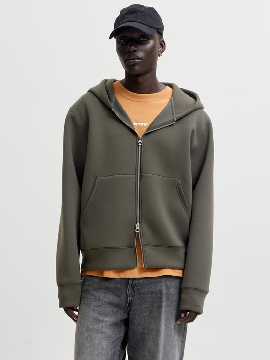 Model draagt een donkergroene hoodie met ritssluiting, oranje T-shirt en spijkerbroek.