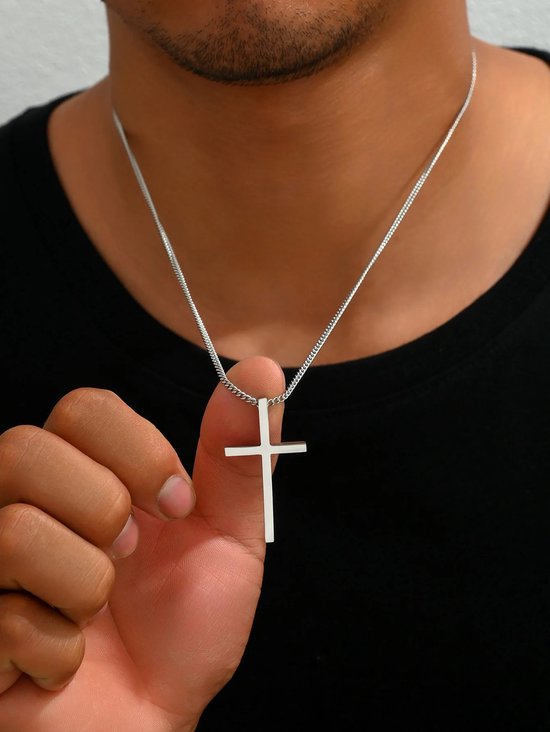 Een man met een zwarte t-shirt houdt een zilverkleurige ketting met kruis-hanger omhoog.