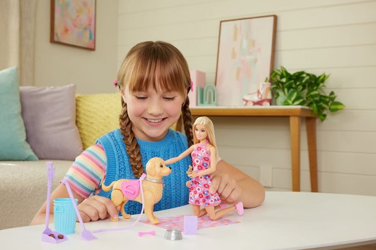 Een meisje speelt met Barbie en een bruine hond met roze tas. Op tafel liggen accessoires en een speelgoedvuilnisbak.