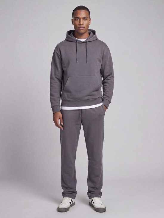 Man draagt een grijze joggingbroek en hoodie met witte t-shirt eronder. Zwarte veters en witte sneakers.