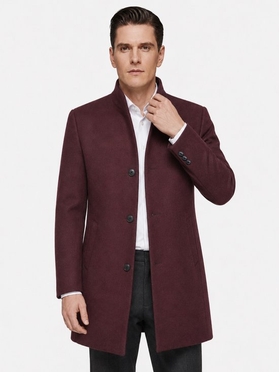 Man in een bordeauxrode wollen jas met opstaande kraag, witte blouse en grijze broek.