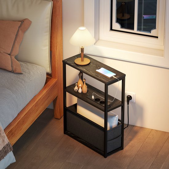 Zwarte bijzettafel naast een bed, met oplaadstation en een tafellamp, in een sfeervol ingerichte slaapkamer.