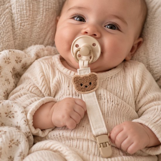 Baby met speen met beige teddybeer speenkoord, die een beige gebreide trui draagt.