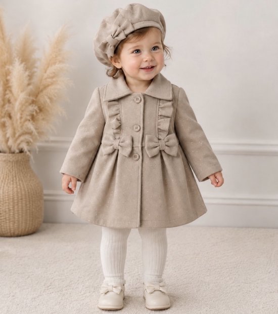 Een baby in een beige jas met ruches, strikjes en glittersteentjes. Ze draagt een bijpassende baret en witte maillot.