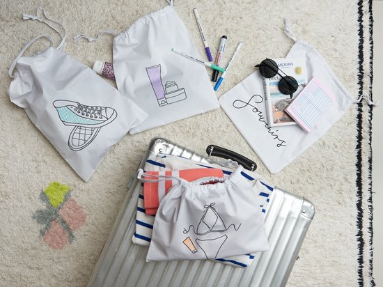 Witte stoffen zakjes met tekeningen van cosmetica, een sneaker en 'Souvenirs'. Een deel van de zakjes ligt op een koffer.