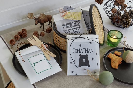 Witte zakjes met kerstdecoratie, waaronder een beer, rendier en dennenappels. Sommige zakjes hebben tekst zoals 'JONATHAN' en 'you can!'.