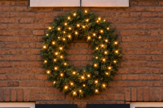 Een klassieke kerstkrans met kunstgroen en 80 warm witte, knipperende LED lampjes, hangend aan een bakstenen muur.