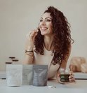 Vrolijke vrouw met krullen met een glas groene drank en drie zakjes supplementen op tafel. Text 'Trusted by women zoals jij'.