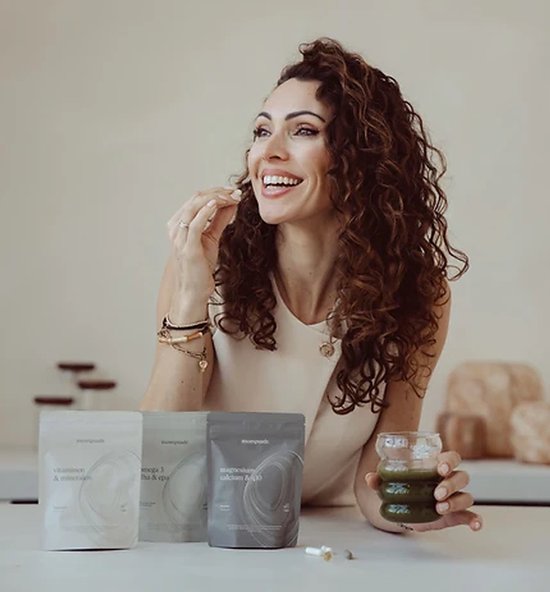 Vrolijke vrouw met krullen met een glas groene drank en drie zakjes supplementen op tafel. Text 'Trusted by women zoals jij'.