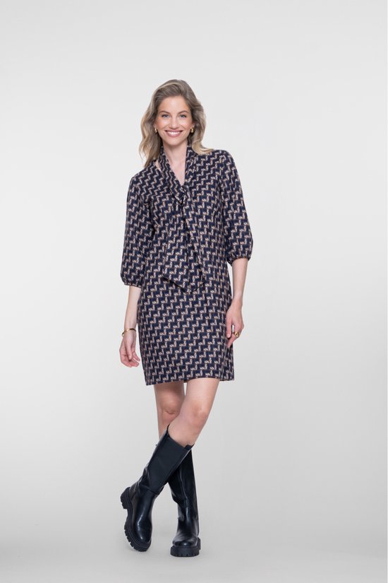 Vrouw met blonde haren, jurk met geometrische print, strikdetail, gouden accessoires en zwarte laarzen.