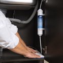 Samsung waterfilter met label, gemonteerd onder een gootsteen, met een witte handdoek en handen zichtbaar.