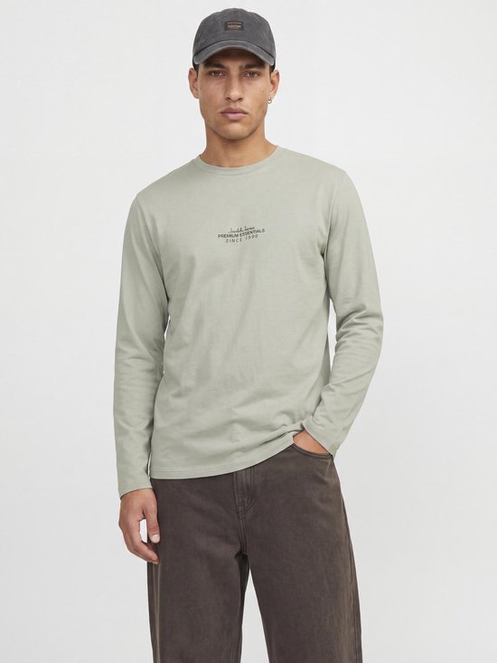 Model draagt een lichtgrijs T-shirt met lange mouwen, gecombineerd met een donkerbruine broek en pet.