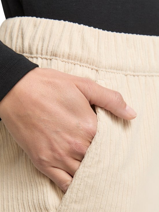 Een hand in de zak van een beige, slanke corduroy broek met verticale ribbels. De broek heeft een elastische tailleband.