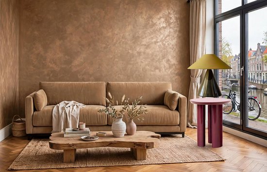Een ruime, taupekleurige bank staat voor een muur met zandkleurige, ruwe textuurverf. Een houten salontafel met bladeren staat voor de bank. Twee vazen, een lamp en een bijzettafel staan naast de bank.