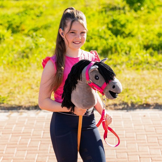 Een meisje met paardenstaart draagt een roze top en donkerblauwe legging, met een grijs hobby horse met roze hoofdstel.