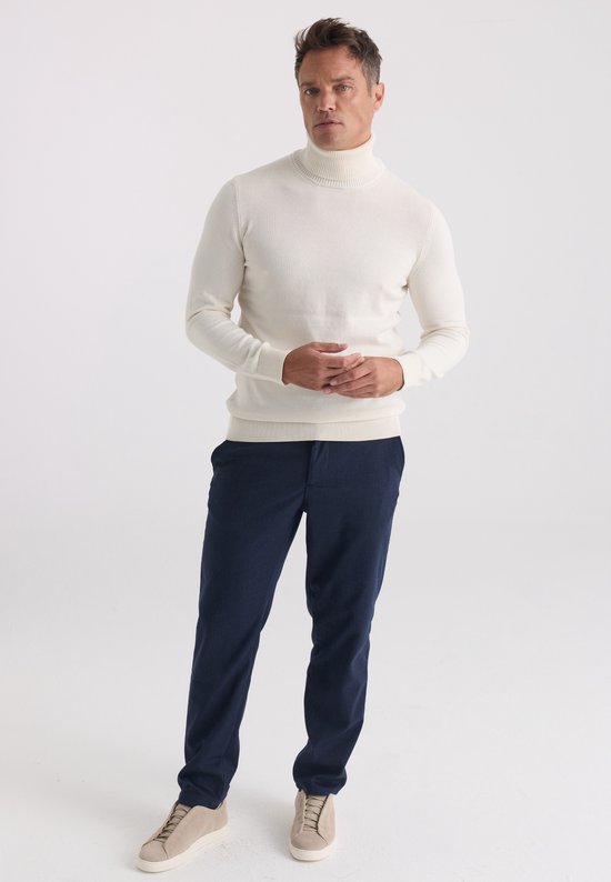 Model draagt een ecru coltrui met lange mouwen, donkerblauwe broek en beige sneakers op een witte achtergrond.