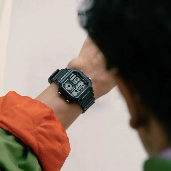 Zwart digitaal Casio-horloge gedragen om de pols door een persoon in een oranje en groen jack.