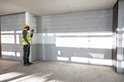 Bouwvakker met veiligheidshelm en neonhesje gebruikt een rode laser-afstandsmeter tegen een grijze muur.