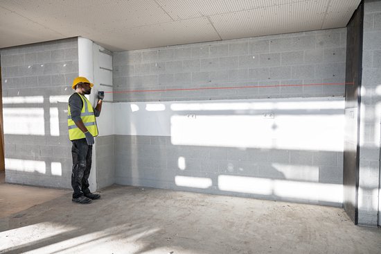 Bouwvakker met veiligheidshelm en neonhesje gebruikt een rode laser-afstandsmeter tegen een grijze muur.