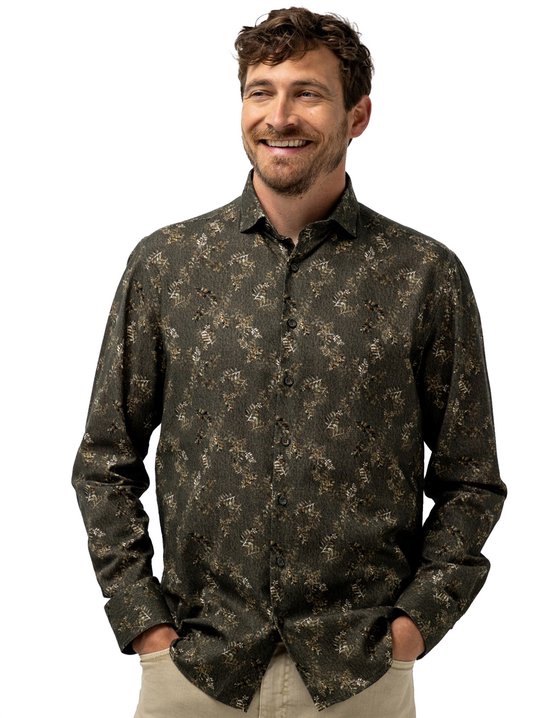 Man in groen overhemd met botanische print en beige broek, handen in de zakken.