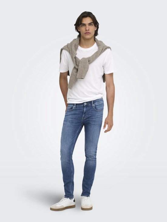 Model in blauwe skinny jeans, wit T-shirt en een beige trui over de schouders.
