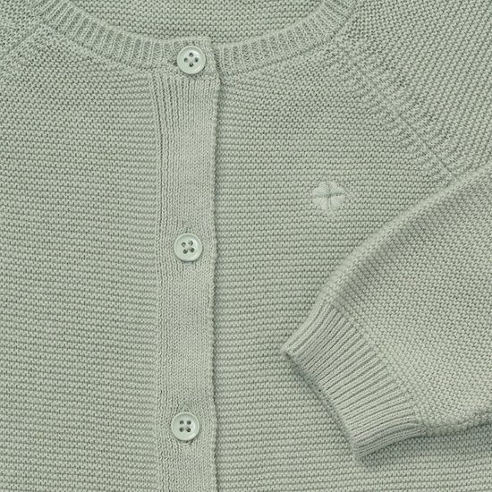 Close-up van de sluiting met knoopjes en een subtiel geborduurd klavertje op een saliegroen gebreid vestje.