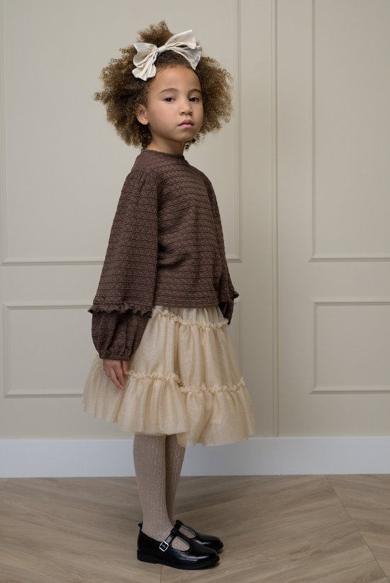 Meisje met krullend haar en strik draagt een bruine longsleeve, beige tule rok en zwarte schoenen.