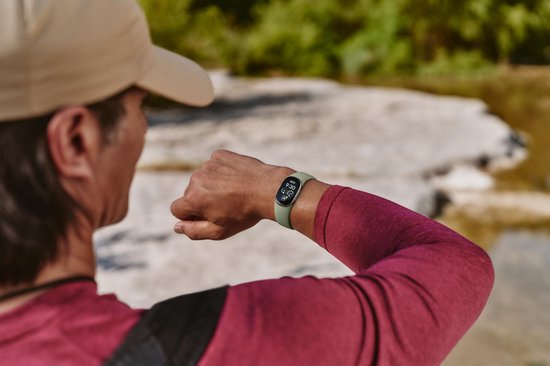 Man met beige pet en bordeauxrode sporttop kijkt naar een olijfgroene Fitbit Edge om zijn pols.