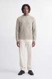 Model in beige grofgebreide trui en witte broek, volledig zicht van voren.