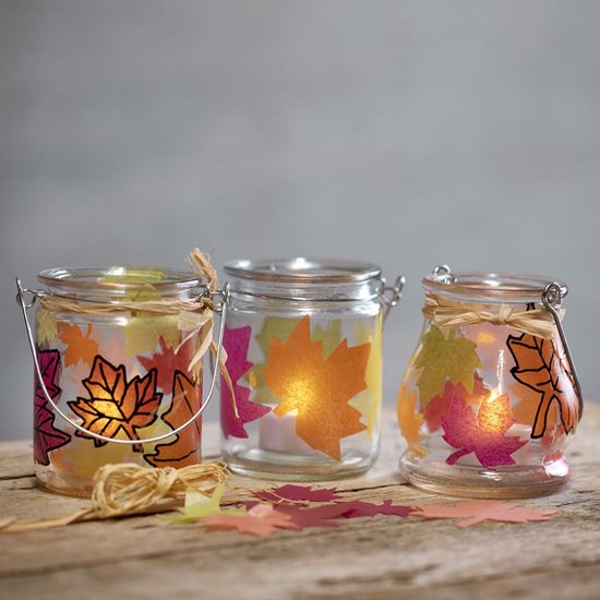 Drie glazen lantaarns versierd met herfstbladeren van vloeipapier en verlicht door theelichtjes.