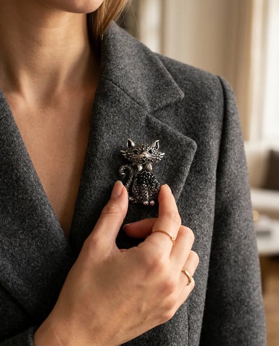 Hand die een zilveren kattenbroche met zwarte kristallen op een grijze blazer speldt.